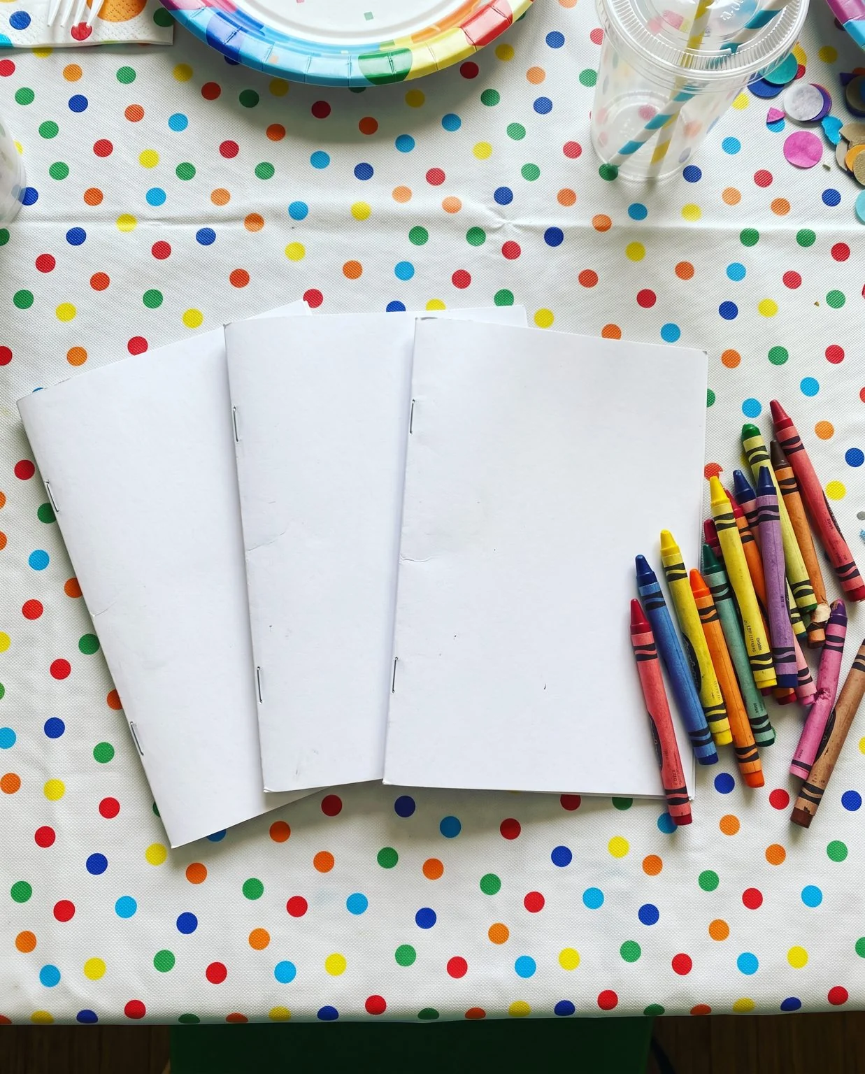 Three stapled half letter coloring booklets with plain covers on a birthday party table next to loose crayons